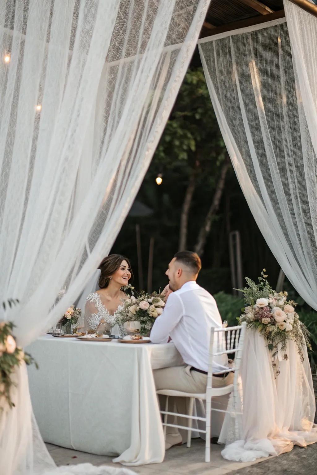 The bride and groom are seated at a table surrounded by sheer veils for an ethereal effect.